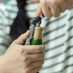 Ręce wyciągają korkociągiem korek od wina z zielonej butelki.