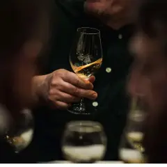 Mężczyzna trzyma kieliszek ze złotym winem muskat. W tle widać inne kieliszki i zamazane sylwetki ludzi.