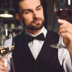 Elegancki mężczyzna w muszce ocenia dwa kieliszki wina. Pokazuje, jak trzymać kieliszek wina, by docenić jego barwę i aromat.