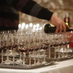 Rzędy kieliszków z czerwonym winem, osoba nalewająca wino z butelki.