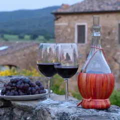Dwa kieliszki czerwonego wina, kiść winogron i butelka w koszyczku, na tle kamiennego muru i krajobrazu regionu Chianti.