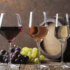 Trzy kieliszki z winem: czerwonym, różowym i białym, obok kiści winogron i beczki. Degustacja francuskiego gatunku wina.