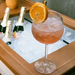 Orzeźwiający drink z prosecco i colą, ozdobiony plasterkiem pomarańczy, obok schłodzonych butelek.