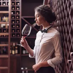 Elegancka sommelierka w białej koszuli i muszce wącha czerwone wino w kieliszku, otoczona regałami pełnymi butelek.
