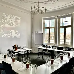 Sala do degustacji win, przygotowana do spotkania klubu wino. Na stołach kieliszki, dzbanki z wodą i butelka wina. Na ekranie logo WSET.