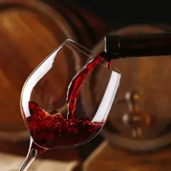 Czerwone wino leje się do kieliszka, a w tle widać drewniane beczki, które opowiadają historię wina.