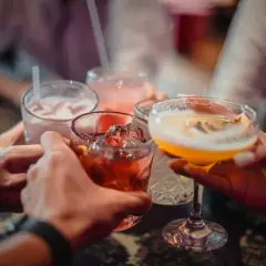 Ręce wznoszą kieliszki z kolorowymi drinkami. Wśród nich widać czerwony koktajl z lodem, różowy napój ze słomką i żółty drink w kieliszku. To idealny moment na toast z dobrym **medoc alkohol**!