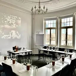Sala do degustacji win, przygotowana do spotkania klubu wino. Na stołach kieliszki, dzbanki z wodą i butelka wina. Na ekranie logo WSET.