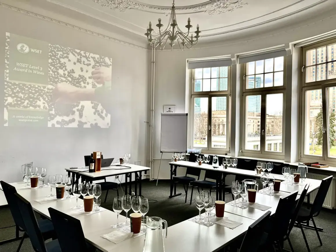 Sala do degustacji win, przygotowana do spotkania klubu wino. Na stołach kieliszki, dzbanki z wodą i butelka wina. Na ekranie logo WSET.