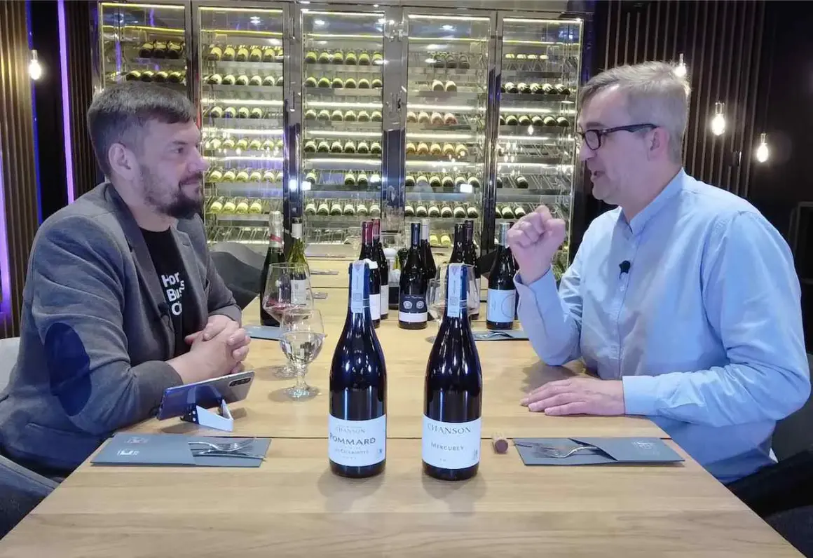 Dwóch mężczyzn przy stole degustuje wino. Na stole stoją butelki, w tym dwie z etykietami sugerującymi pinot noir.