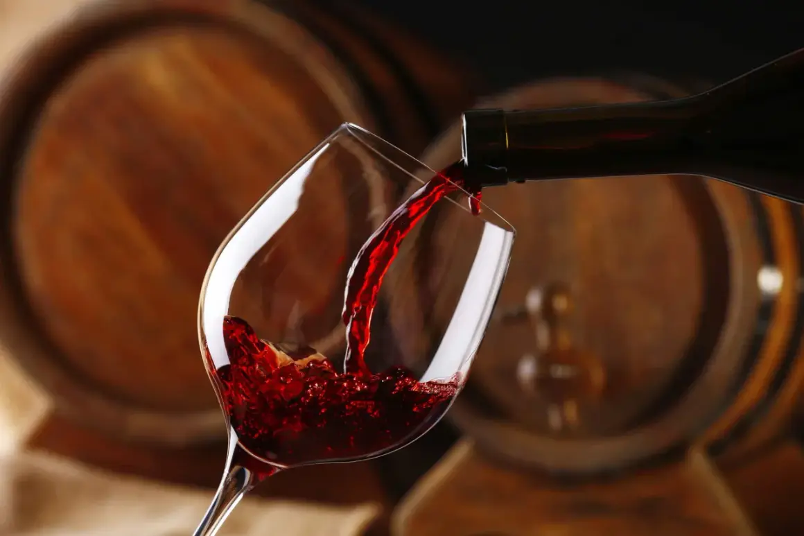 Czerwone wino leje się do kieliszka, a w tle widać drewniane beczki, które opowiadają historię wina.