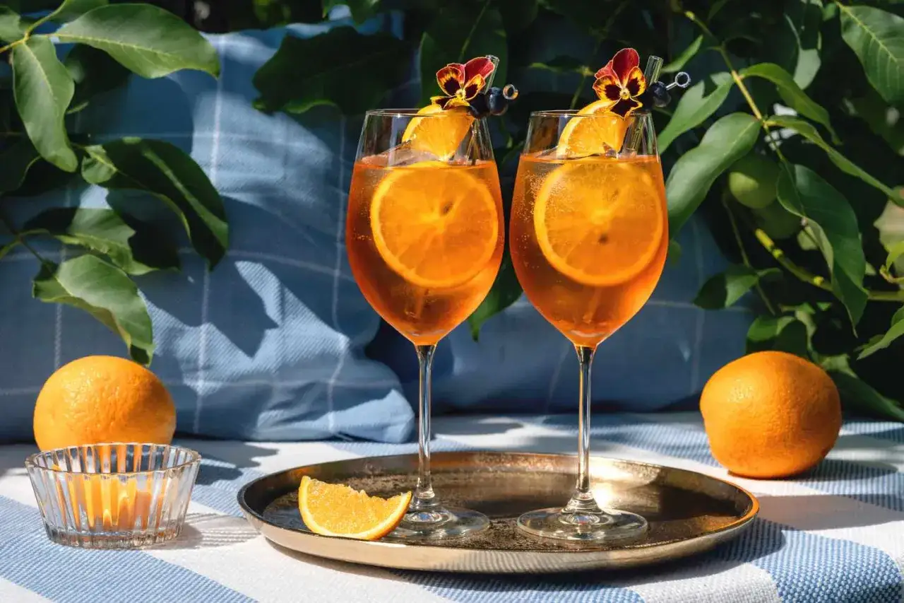 Dwa orzeźwiające drinki Aperol Spritz, ozdobione plastrami pomarańczy i kwiatami, na tle zieleni. Idealne na letnie popołudnie.