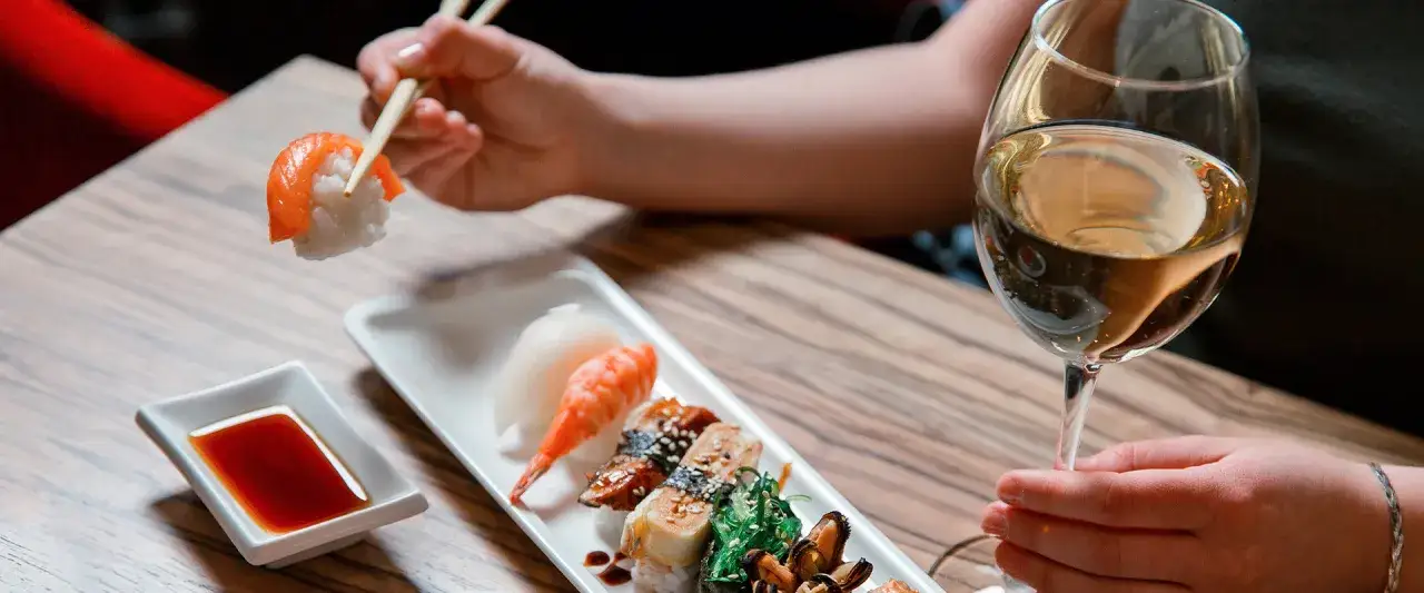 Ręka trzyma pałeczkami kawałek sushi z łososiem. Obok kieliszek białego wina do sushi i talerz z różnymi rodzajami sushi.